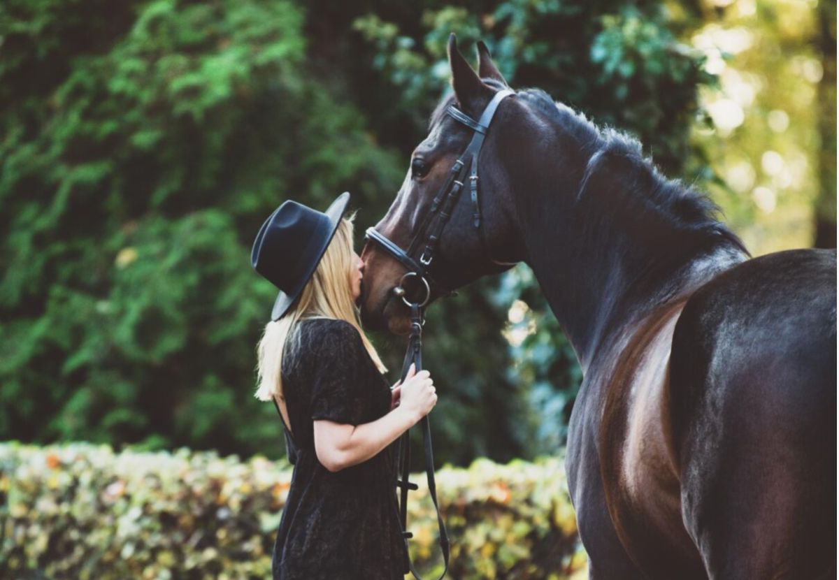 Ung kvinde med sort hat kysser stor mørkebrun hest på mulen.