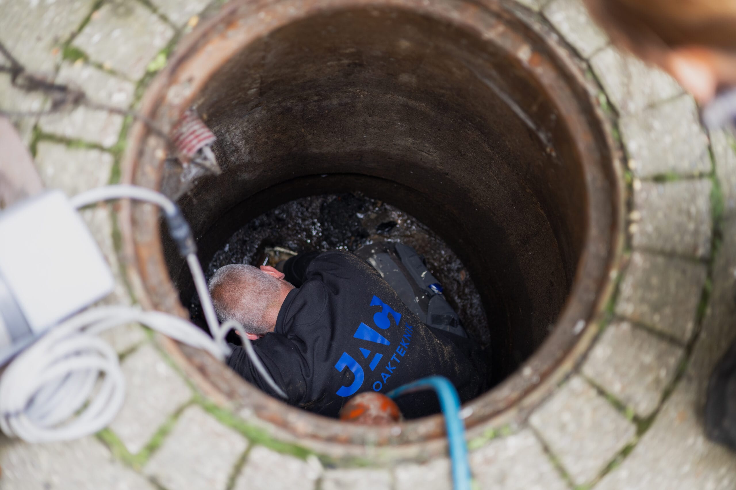Medarbejder fra JAC Kloakservice arbejder i en kloakbrønd for at inspicere og vedligeholde kloaksystemet. Billedet viser praktisk kloakarbejde, der kan forebygge tilstopninger og sikre fri gennemstrømning.