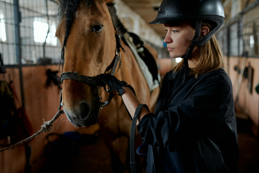 Kvinde med ridehjelm står sammen med en brun hest i stalden.